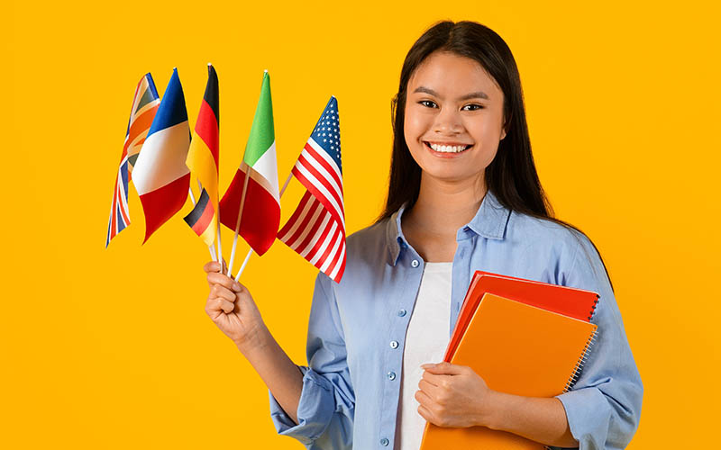 Smiling student holding international flags and notebooks against a bright yellow background.
