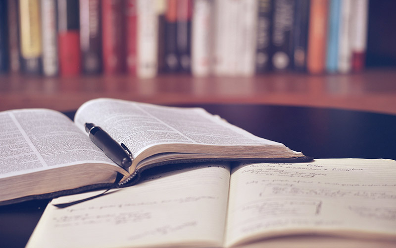 Open book with pen beside handwritten notes, with a blurred bookshelf background for study setting.