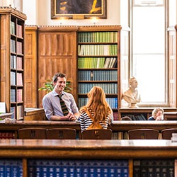 Man and woman talking at a library desk surrounded by bookshelves, busts, and large windows.