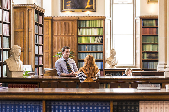 Man and woman talking at a library desk surrounded by bookshelves, busts, and large windows.