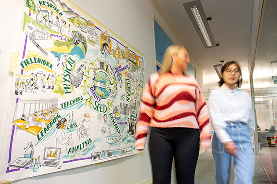 Two women walk past a colourful illustrated geography-themed poster on a hallway wall in an academic building.