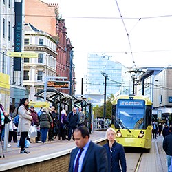 Crowd waits at Market Street tram stop in Manchester as a yellow tram approaches on a busy shopping street.