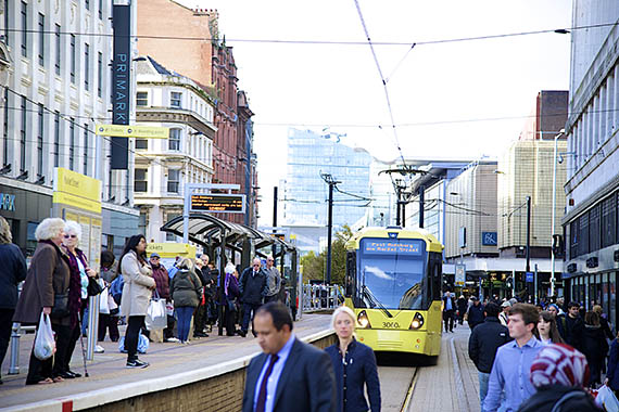 Crowd waits at Market Street tram stop in Manchester as a yellow tram approaches on a busy shopping street.