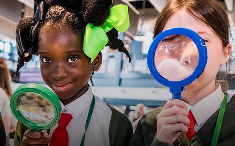 Three schoolchildren in uniform smiling and looking through colourful magnifying glasses indoors.
