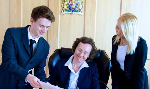 Three legal workers review a document in a formal meeting room as a seated woman smiles while two colleagues stand beside her discussing paperwork.