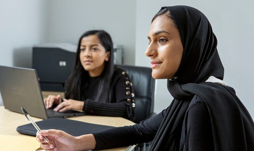 Woman wearing a black hijab holds a pen during a meeting while a colleague works on a laptop in the background of a bright, modern office.