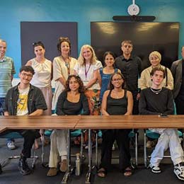 Diverse group of adults posing together in a classroom, seated and standing behind a table, smiling at the camera.
