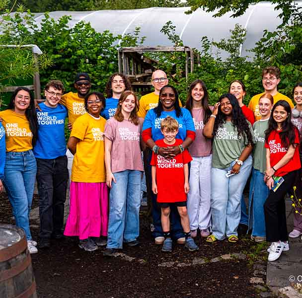 Diverse group in colourful "One World Together" shirts smiling outside near greenhouse and plants.