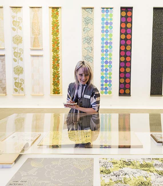 Woman at table with colourful textile panels hanging vertically on the wall behind her.