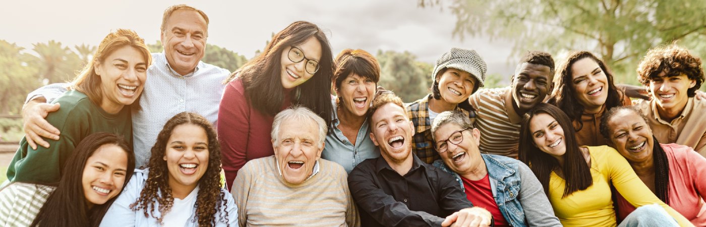 Group of diverse adults of various ages sitting on grass, laughing together with arms around each other in a sunny park.