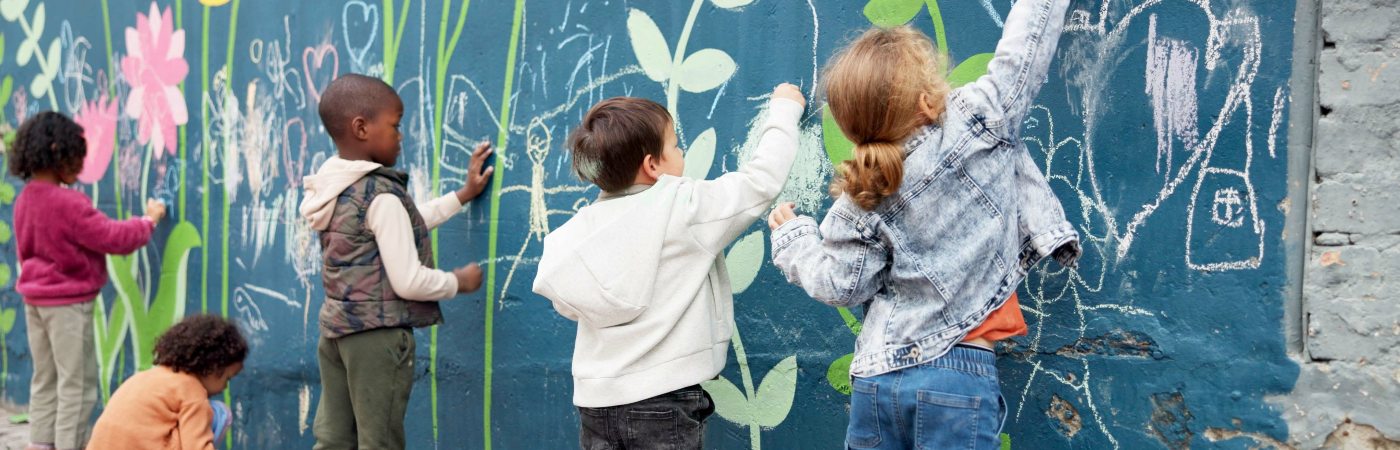 Children drawing with chalk on a colourful mural wall with flowers and doodles, standing and kneeling on a sidewalk outdoors.