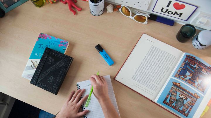 Someone writing on a desk which is full of books and study materials