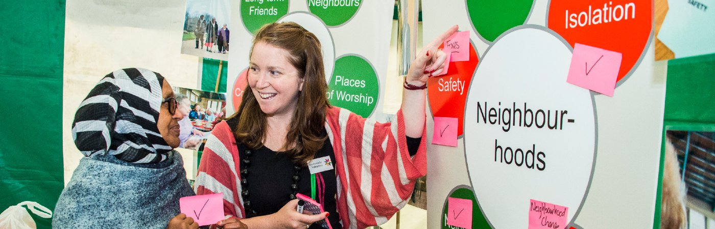 Two woman smiling and pointing at a display board featuring words such as neighbourhoods and friendliness.