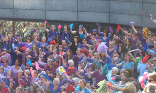 Large, colourful crowd waving bright scarves and fabric in the air during a lively outdoor gathering.