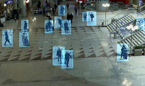 Crowded transit hall with people tracked by blue facial recognition boxes near escalators.