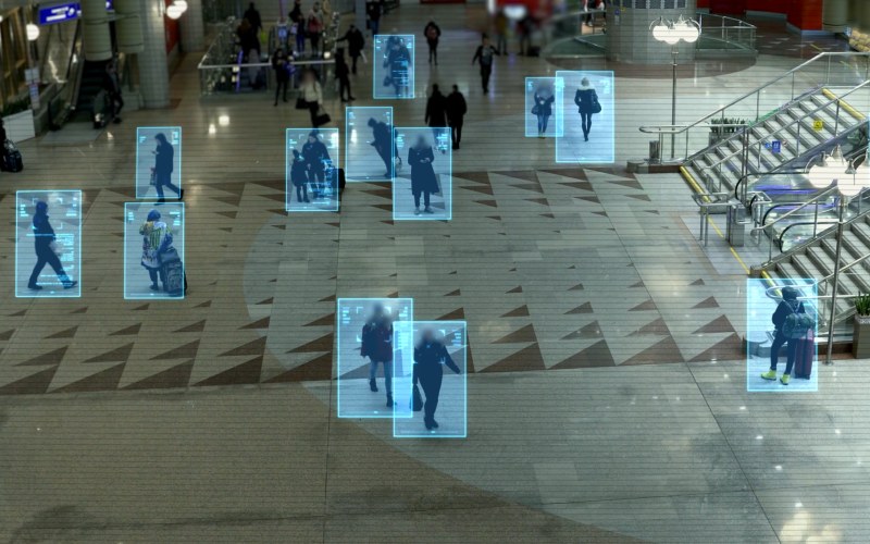 Crowded transit hall with people tracked by blue facial recognition boxes near escalators.