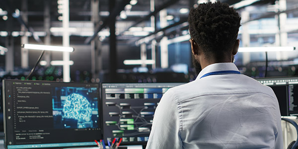 Back view of engineer at dual monitors showing code and AI brain graphic in modern data centre.