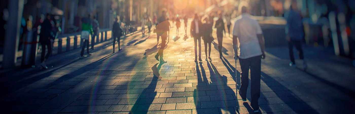 A blurred image of people walking down the pavement in sunny conditions.