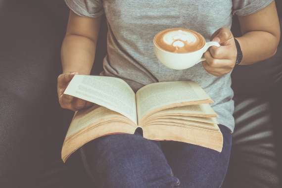 A woman reading a book in her lap while holding a cup of coffee 
