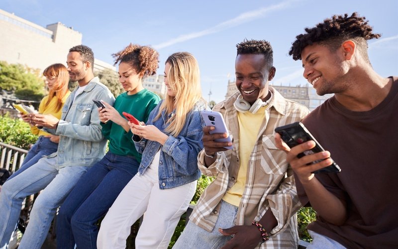 A diverse group of young adults sitting outdoors, looking at their smartphones together.