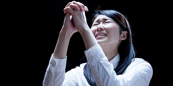 Woman in white shirt clasping hands and looking up with a pained, pleading expression in the dark.