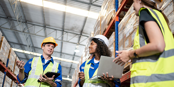Warehouse workers discuss inventory using a tablet and laptop between storage racks.