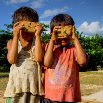 Two young boys holding rocks over their faces like masks while standing outdoors on a sunny day.
