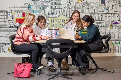 Four women sitting around a table working on laptops, with a colourful city mural in the background.