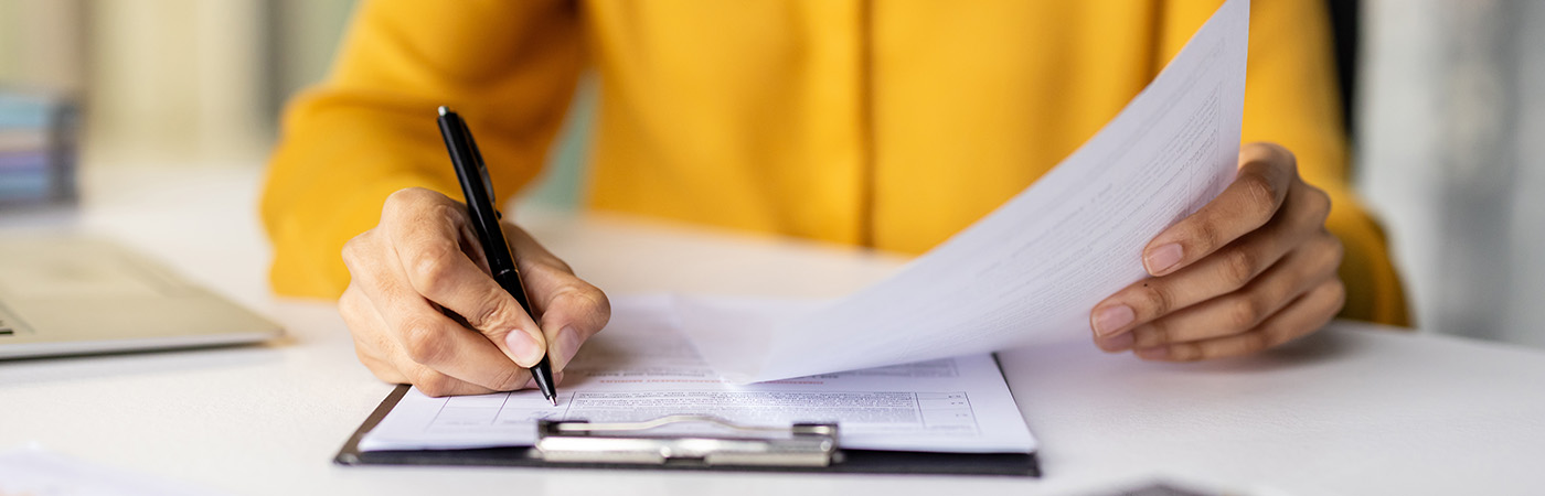 Person reviewing and signing documents at a desk with paperwork and a tablet.