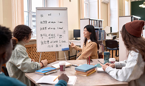 Side view of smiling Asian woman as female teacher teaching Chinese language class to students by whiteboard in library.
