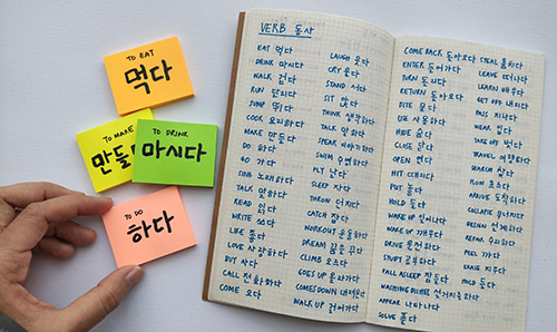 Basic Korean words and verbs written in sticky notes and notebook.